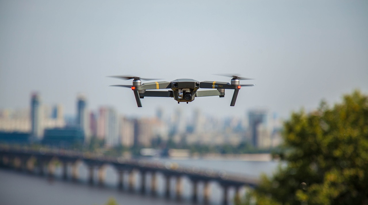 A drone with a camera is in the air with a city in the background.