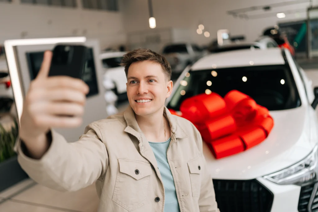 Happy car buyer taking a selfie in front of their newly purchased vehicle at a dealership.